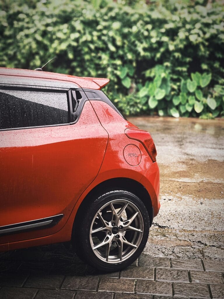 Close-up of a red car parked in the rain with lush green foliage in the background.